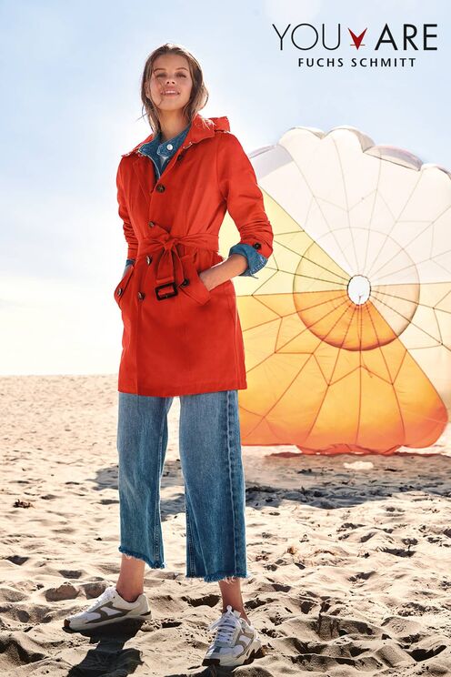 Blondes Model am Strand trägt eine weite knöchellange Jeans sowie einen auffällig roten Mantel der Modemarke Fuchs Schmitt